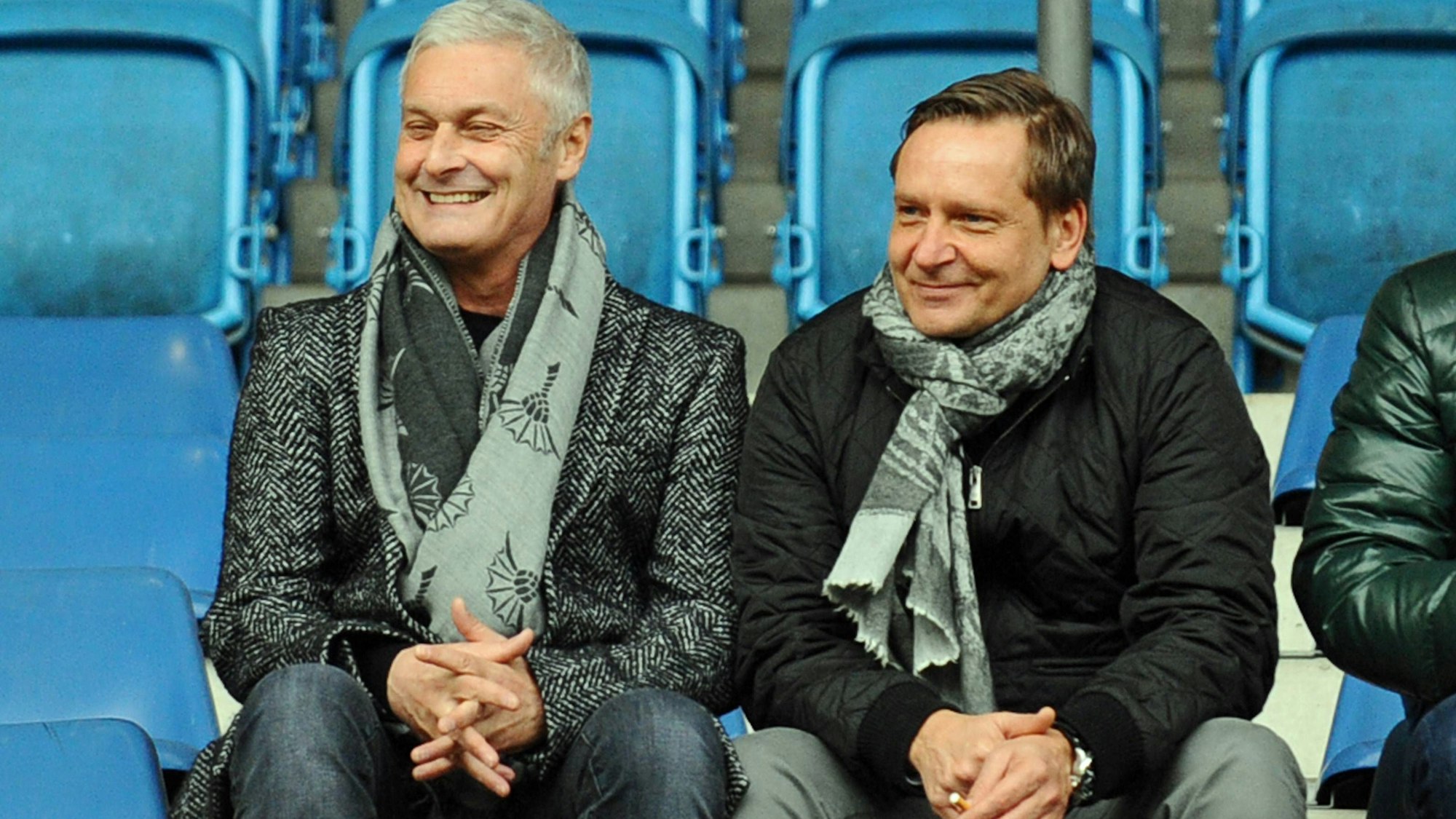 Armin Veh und Horst Heldt, zwei ehemalige Geschäftsführer des 1. FC Köln, sitzen nebeneinander auf einer Tribüne und sehen sich entspannt lächelnd ein Fußballspiel an.