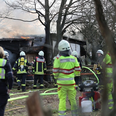 Feuerwehrleute, Sanitäter und Polizeibeamte stehen während eines Brands an einer Scheune im Kölner Stadtteil Rodenkirchen am Samstag, 28. Januar 2023. Bei dem Feuer ist eine Person tot aufgefunden worden.