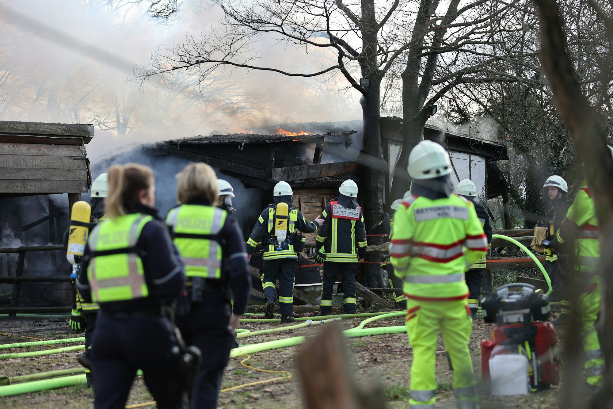 Feuerwehrleute, Sanitäter und Polizeibeamte stehen während eines Brands an einer Scheune im Kölner Stadtteil Rodenkirchen am Samstag, 29. Januar 2023. Bei dem Feuer ist eine Person tot aufgefunden worden.