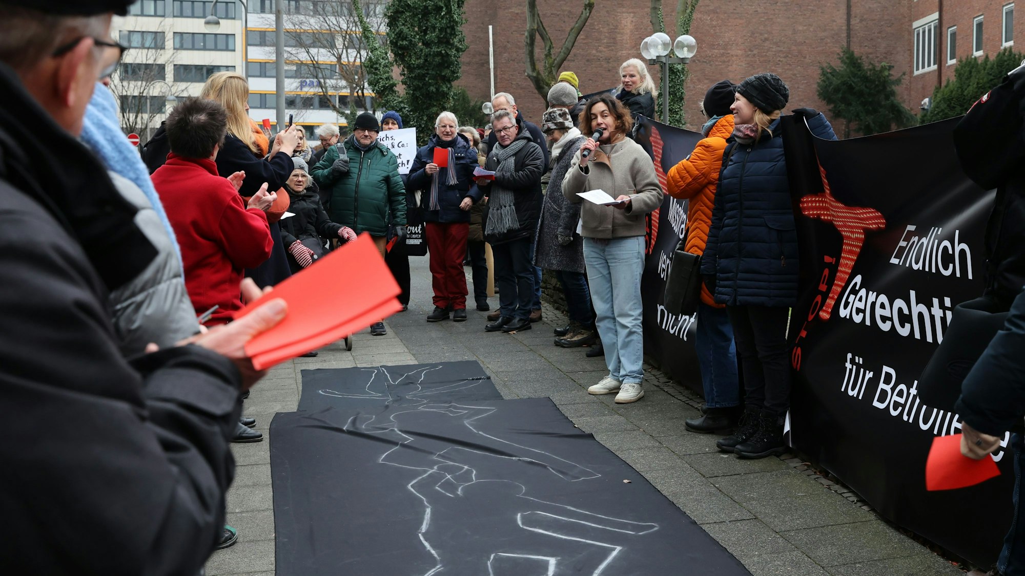 Etwa 70 Menschen kamen zu der Kundgebung vor dem Erzbischöflichen Palais in Köln. Die Teilnehmenden halten rote Karten und Transparente. Auf dem Boden ist ein schwarzes Tuch ausgebreitet, auf dem mit Kreide die Umrisse liegender Menschen nachgezeichnet sind.