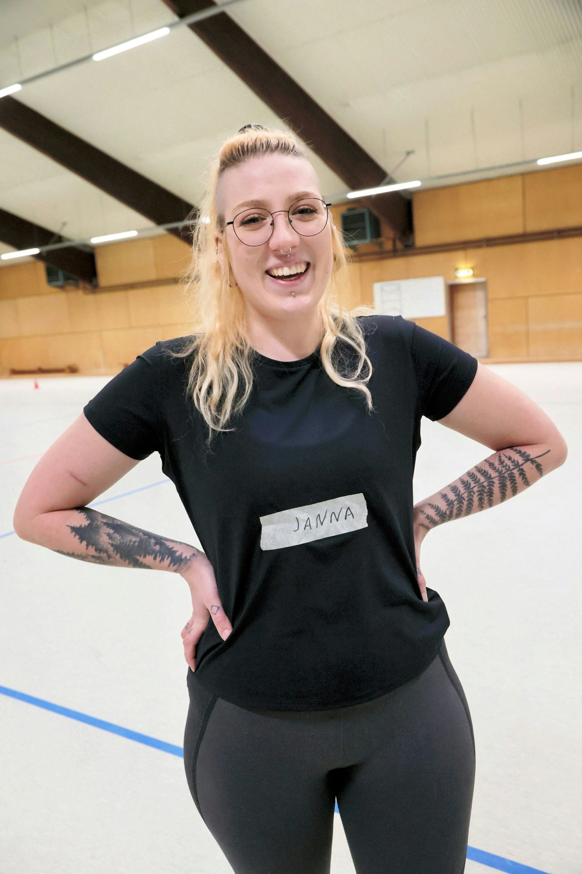 Die 26-Jährige trägt dunkle Sportkleidung, hat Tattoos an den Unterarmen und blonde Haare mit Side Cuts.