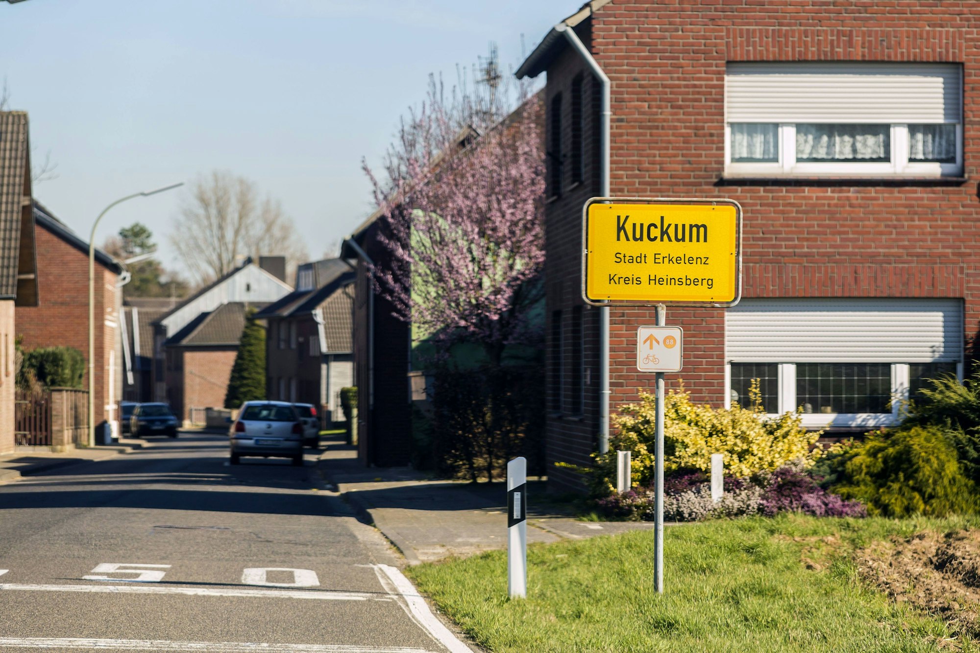 Das Ortsschild von Kuckum steht am Straßenrand, dahinter sind Wohnhäuser und einige geparkte Autos zu sehen. Ein Baum blüht rosa, ein Busch gelb im Vordergrund.
