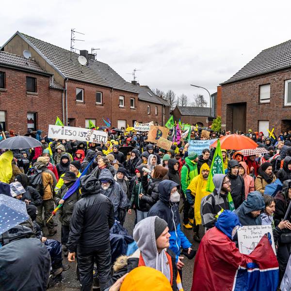 Das Bild zeigt Menschen bei einer Demo gegen den Abriss des Braunkohle Dorf Lützerath im Ort Keyenberg. Sie schwenken Fahnen und halten Schilder hoch, viele tragen Regenschutz und Schirme - das Wetter ist trüb.