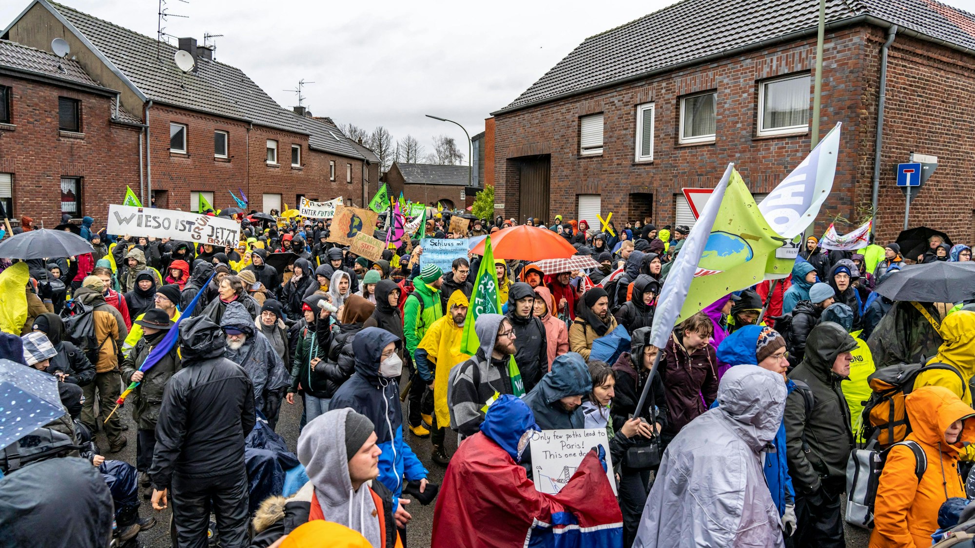 Das Bild zeigt Menschen bei einer Demo gegen den Abriss des Braunkohle Dorf Lützerath im Ort Keyenberg. Sie schwenken Fahnen und halten Schilder hoch, viele tragen Regenschutz und Schirme - das Wetter ist trüb.
