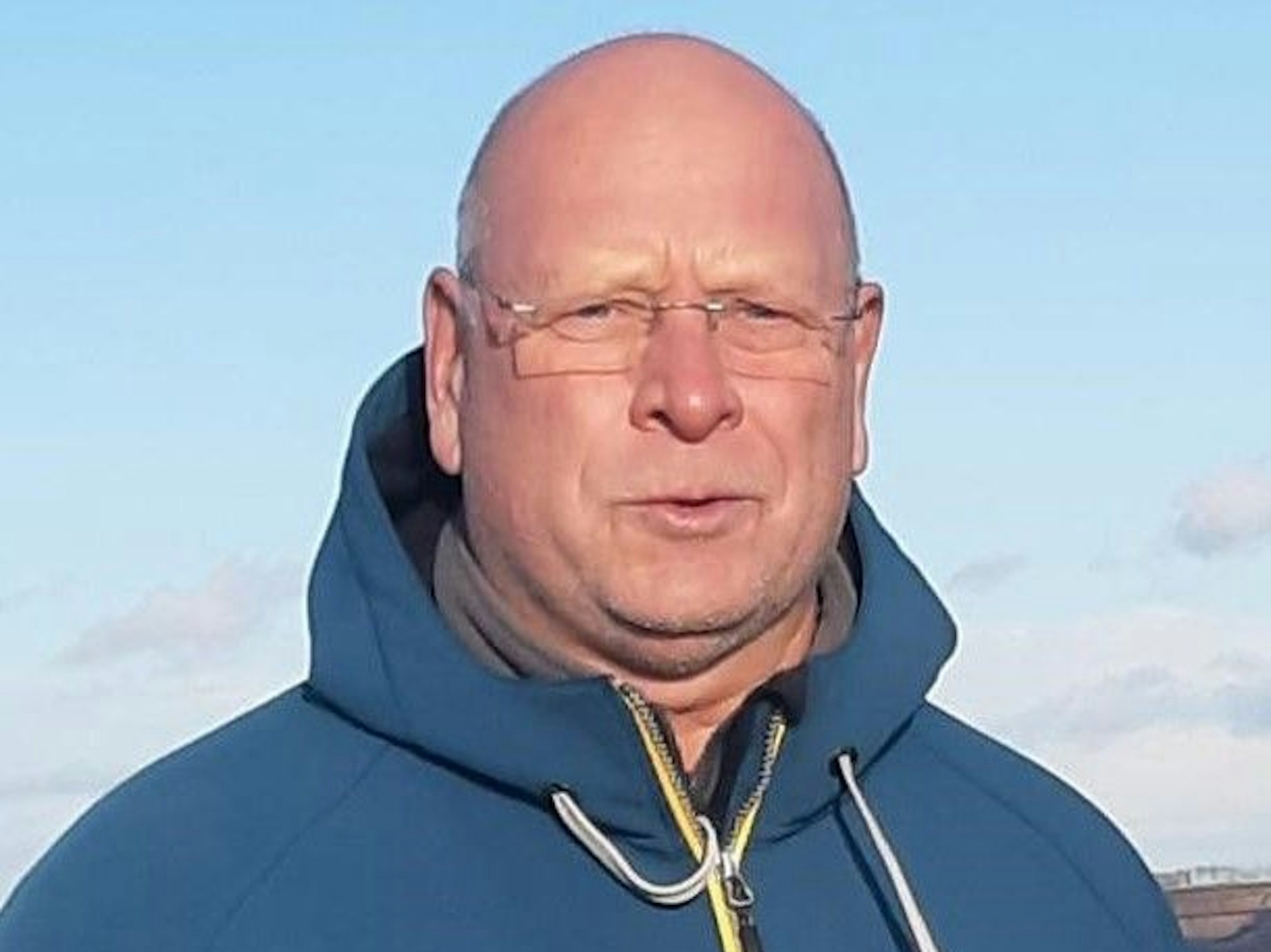 Oliver Kanneberg blickt freundlich in die Kamera. Er trägt eine randlose Brille und eine blaue Kapuzenjacke. Hinter ihm ist ein Stück Tagebau zu sehen unter blauem Himmel.