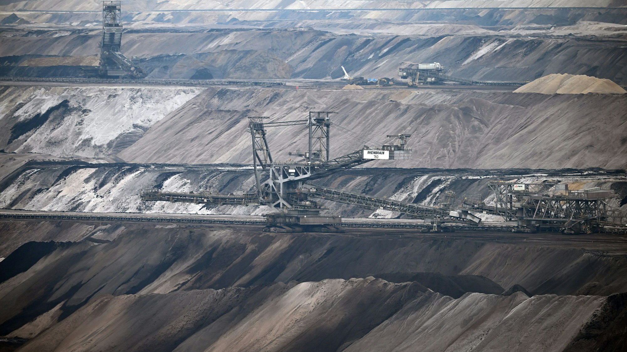 Bergbaumaschinen arbeiten im Tagebau Garzweiler. Weine wüstenartige grau-braune Terrassenlandschaft durchzieht das Bild. In der Mitte sind riesige Maschinen zu sehen.