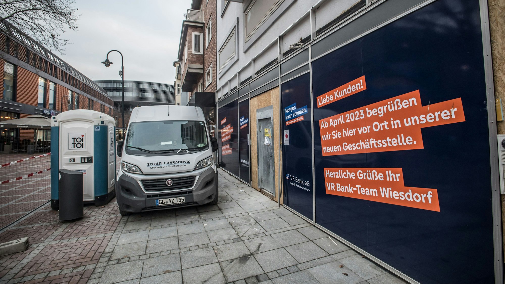 Vor einem Ladenlokal stehen ein Baufahrzeug und eine Baustellentoilette. An der Fassade wird angekündigt, dass in dem Ladenlokal die VR-Bank einzieht.
