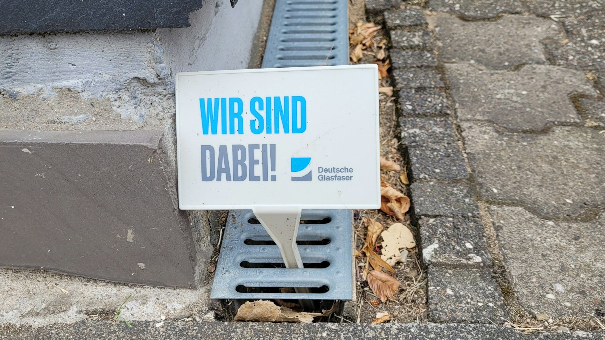Ein Schild der Deutschen Glasfaser mit der Aufschrift „Wir sind dabei“ steckt an Gehwegrand in Kürten.