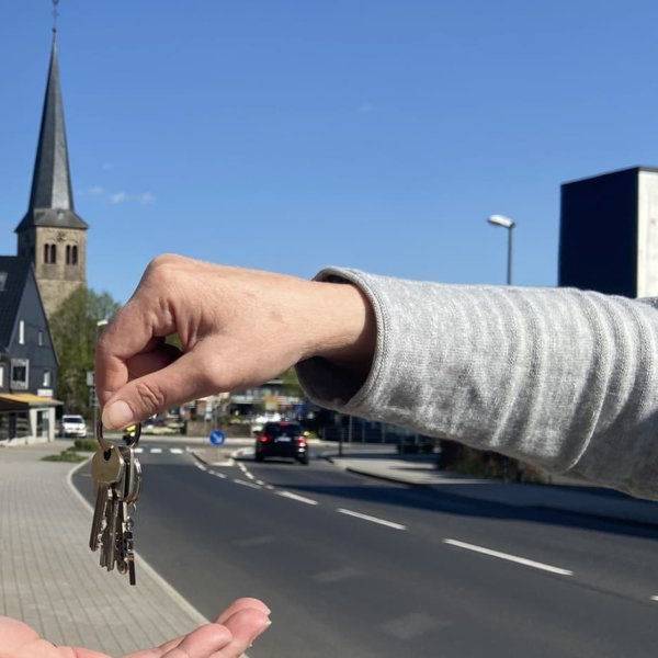 Symbolbild einer Schlüsselübergabe von Hand zu Hand, im Hintergrund eine Dorfkulisse mit Kirchturm.