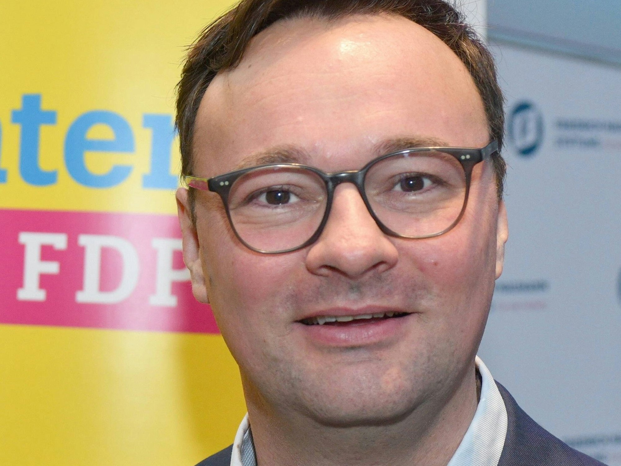 Oliver Luksic ist im Porträt zu sehen vor einem Wahlplakat seiner Partei, der FDP. Er ist Staatssekretär im Bundesverkehrsministerium von Minister Volker Wissing.