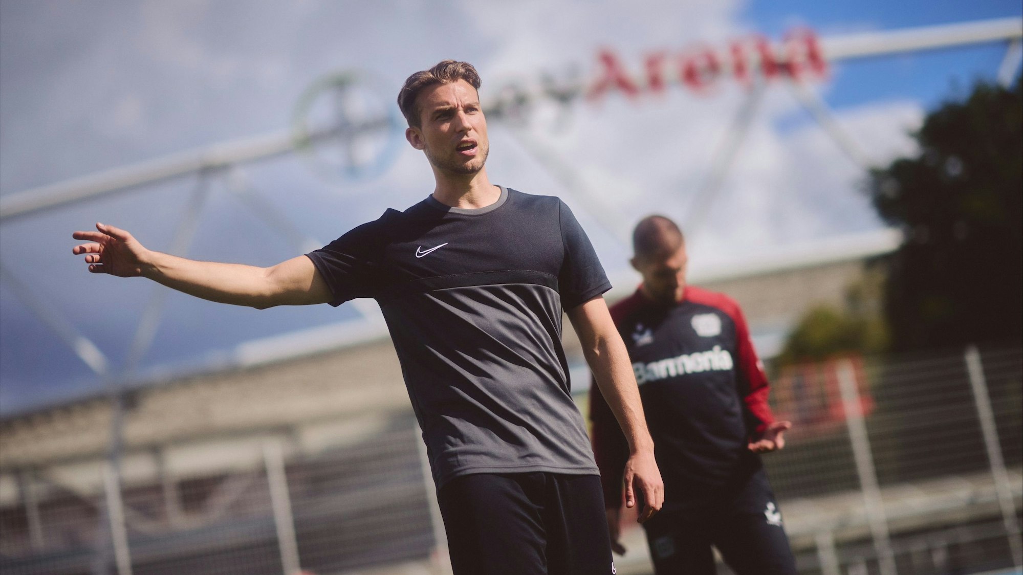 Dennis Eck (vorne) steht vor der BayArena und gibt mit erhobenem Arm Anweisungen, Fußball-Profi Robert Andrich von Bayer 04 Leverkusen schaut auf seine linke Hand.