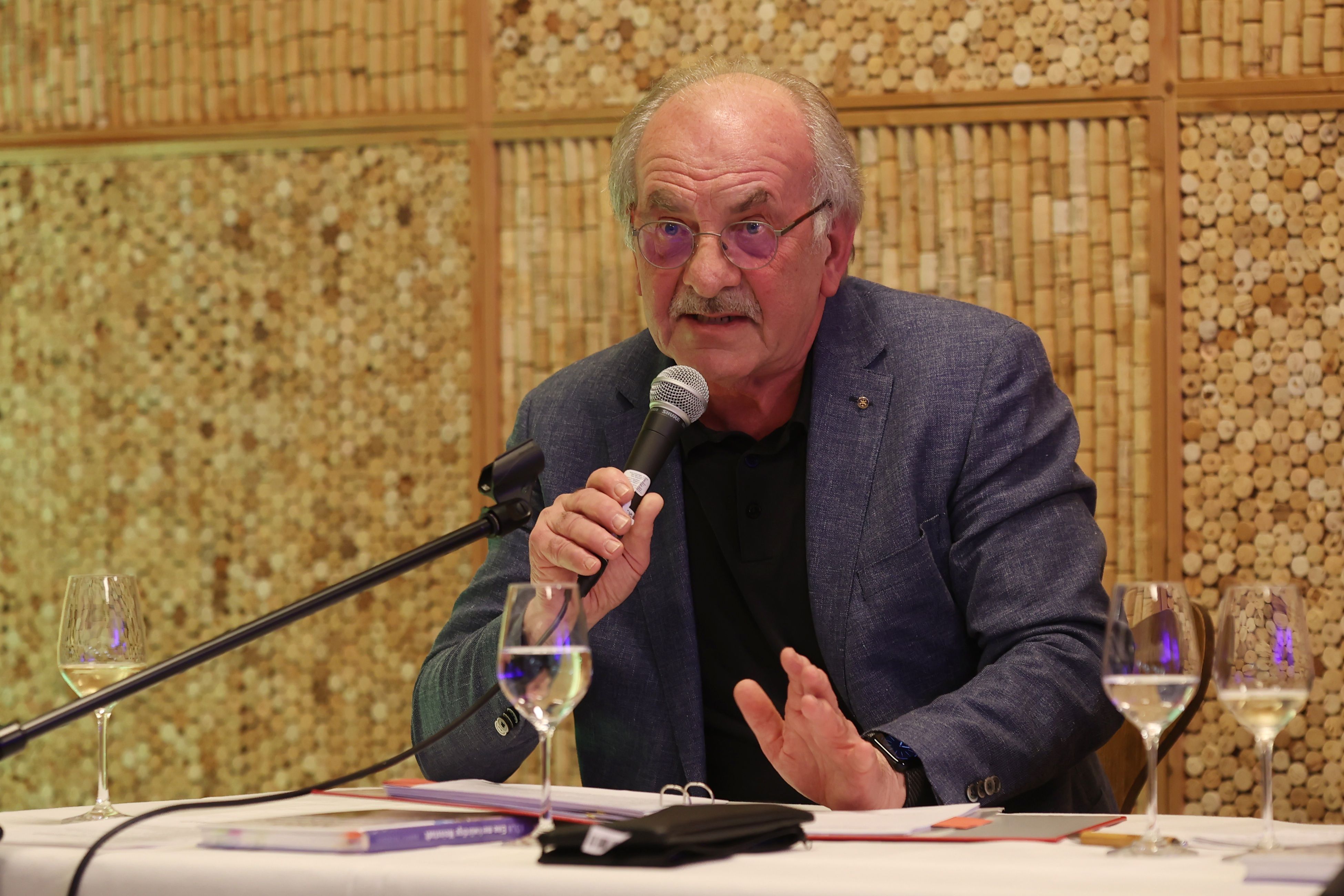 Rudolf Nickenig im Wein-Depot mit einem Mikrofon in der Hand und Weingläsern vor sich stehend.