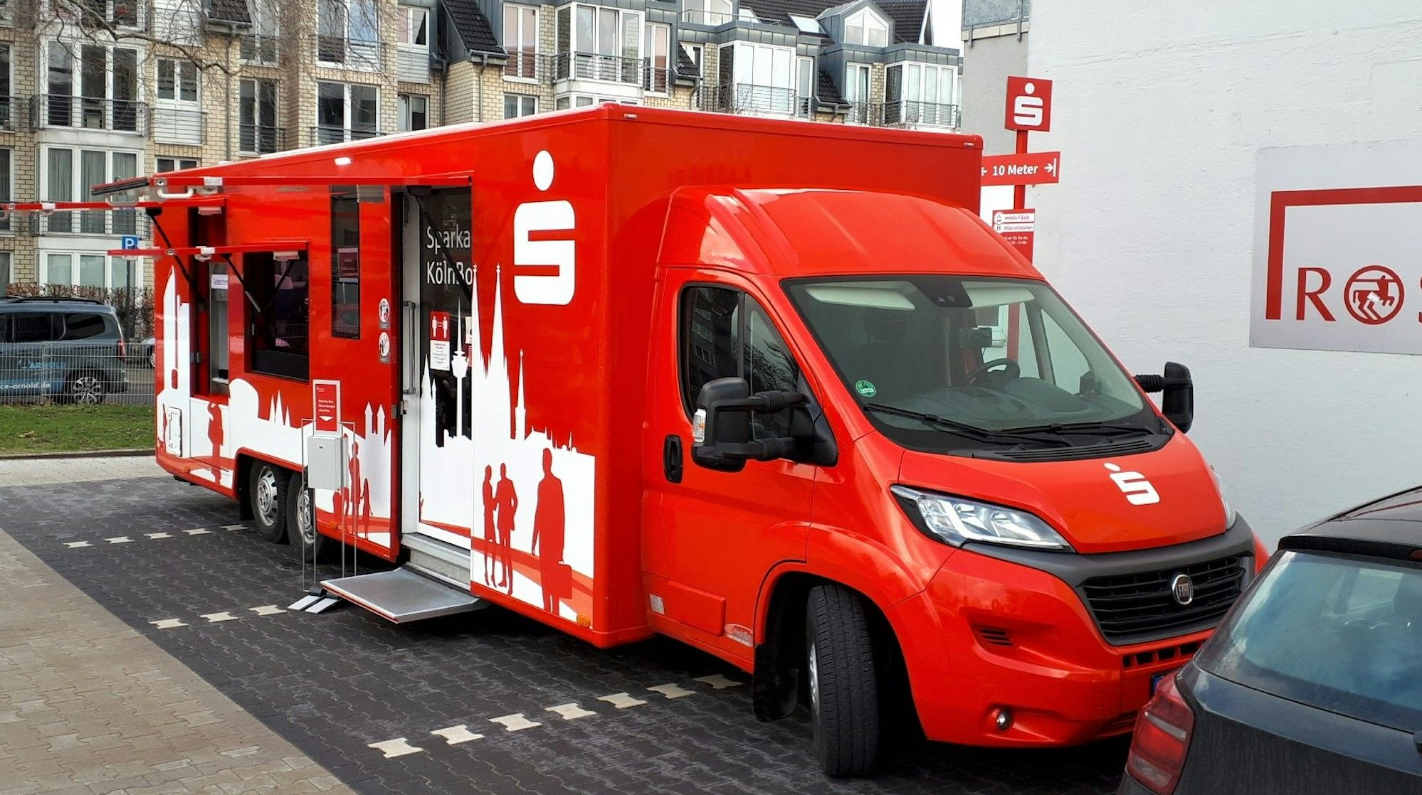 Auf einem Drogeriemarktplatz steht ein großer rot-weißer Filialbus der Sparkasse Köln-Bonn. Neben einem Geldautomaten ist ein überdachter Einstieg zum Innenraum zu sehen. Die Busse sollen die 22 Filialen ersetzen, die die Sparkasse Köln-Bonn schließt.