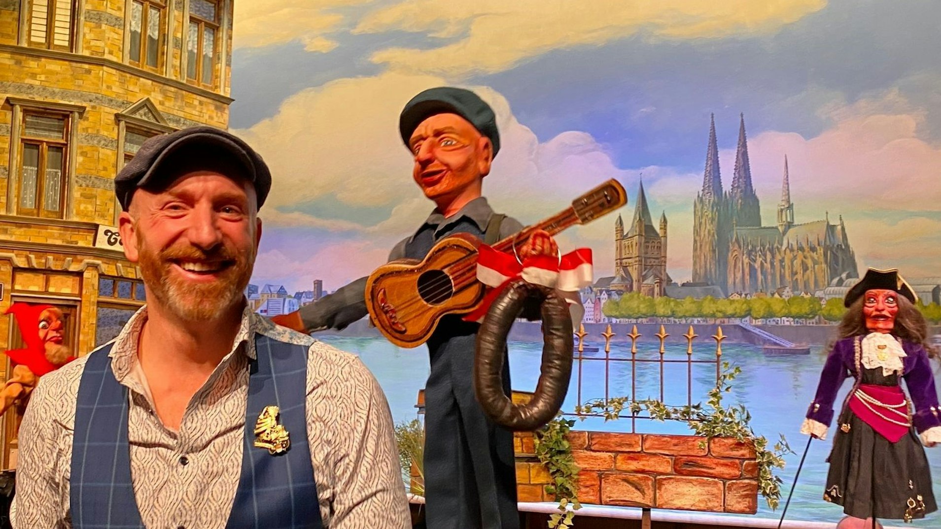 Der Kölner Redner Martin Schopps steht neben seiner Stockpuppe in der Kulisse der Hänneschen-Puppensitzung. Diese zeigt das Kölner Panorama von Deutz aus.
