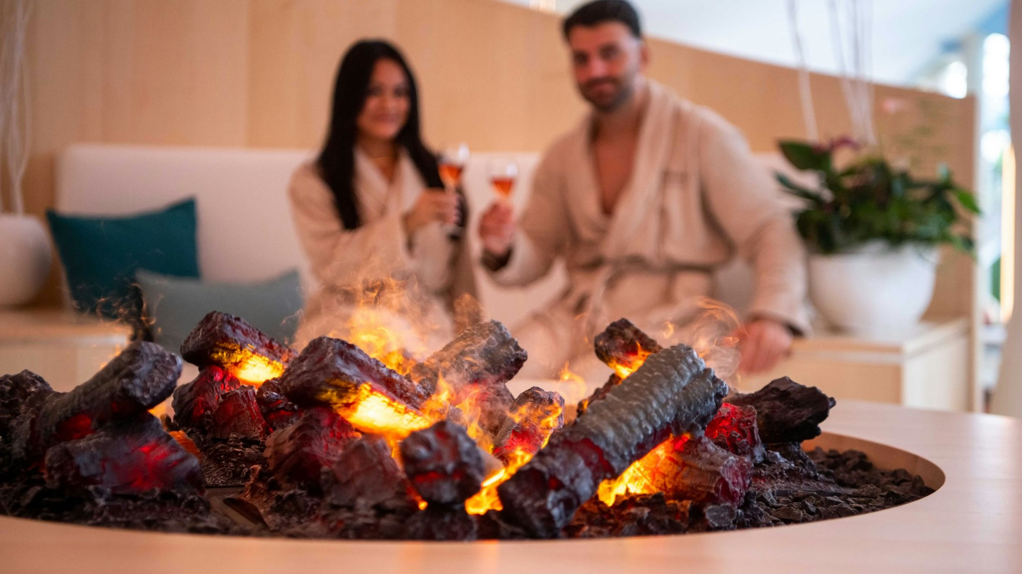 Ein Mann und eine Frau sitzen mit Weingläsern auf einem Sofa, im Vordergrund ist eine Feuerstelle.