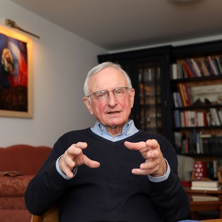 Der katholische Seelsorger Heinz-Peter Janßen sitzt im Wohnzimmer seines Hauses in Bergisch Gladbach-Herkenrath vor einer Bücherwand und spricht.