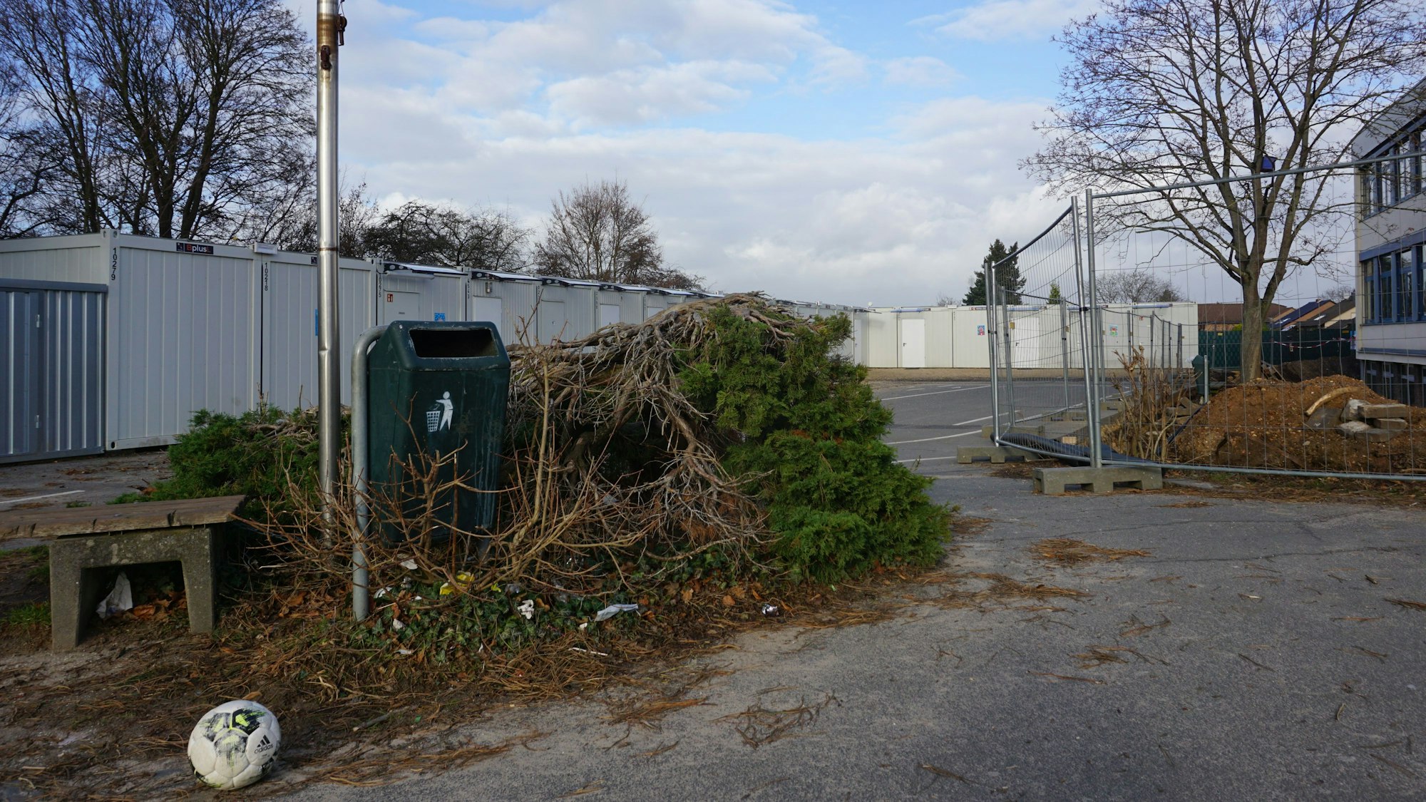 Auf einem trist aussehenden Schulhof stehen Baucontainer und Bauzäune. Dort liegen Schutt, ein alter Fußball und abgeholzte Bäume.