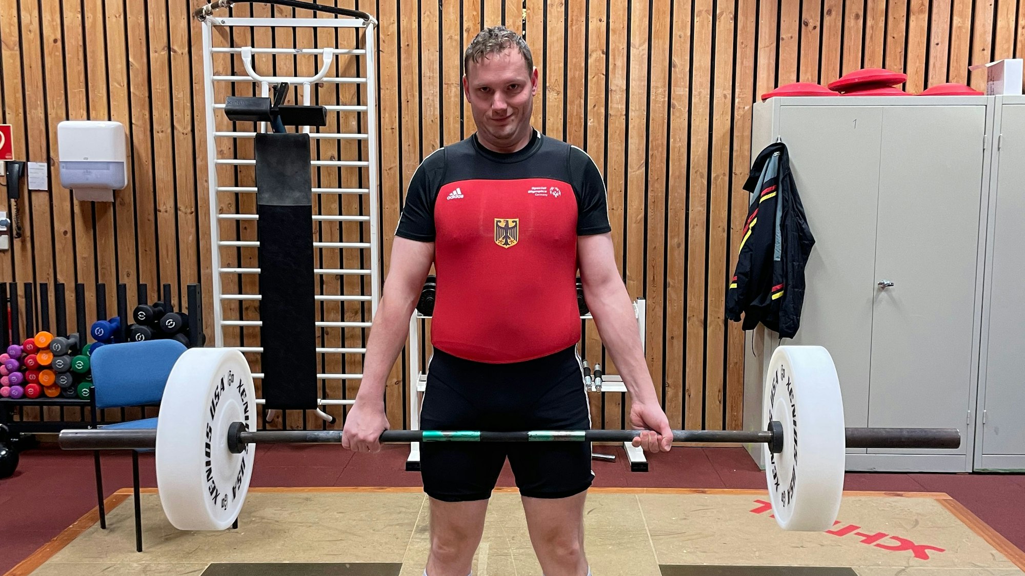 Kai Krüger hält eine Langhantel in beiden Händen. Er trägt rot-schwarze Kraftsportkleidung.