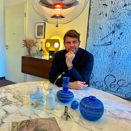 Ein Mann mit braunem Haar und Bart sitzt an einem Tisch mit Marmorplatte. Im Hintergrund ist ein abstraktes Gemälde zu sehen.