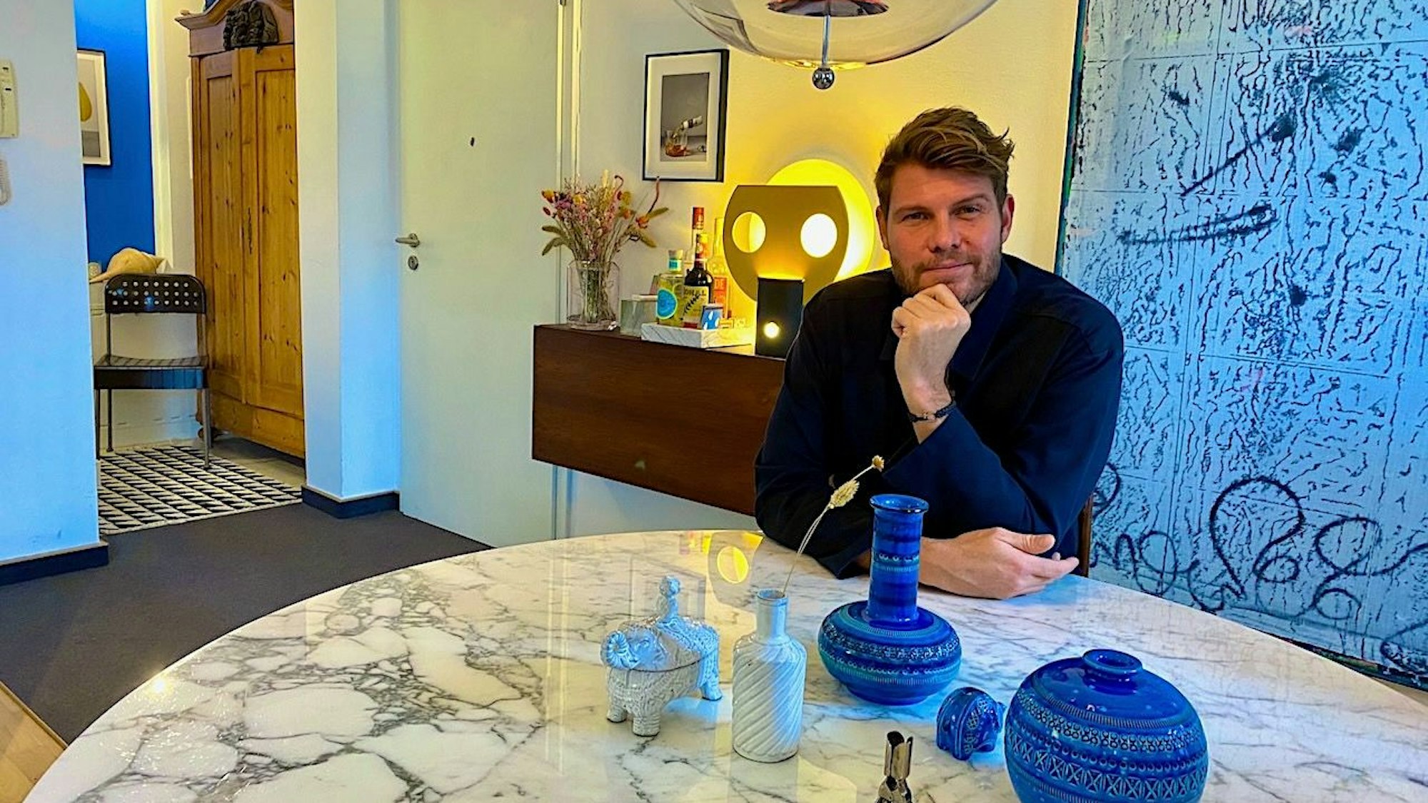 Ein Mann mit braunem Haar und Bart sitzt an einem Tisch mit Marmorplatte. Im Hintergrund ist ein abstraktes Gemälde zu sehen.