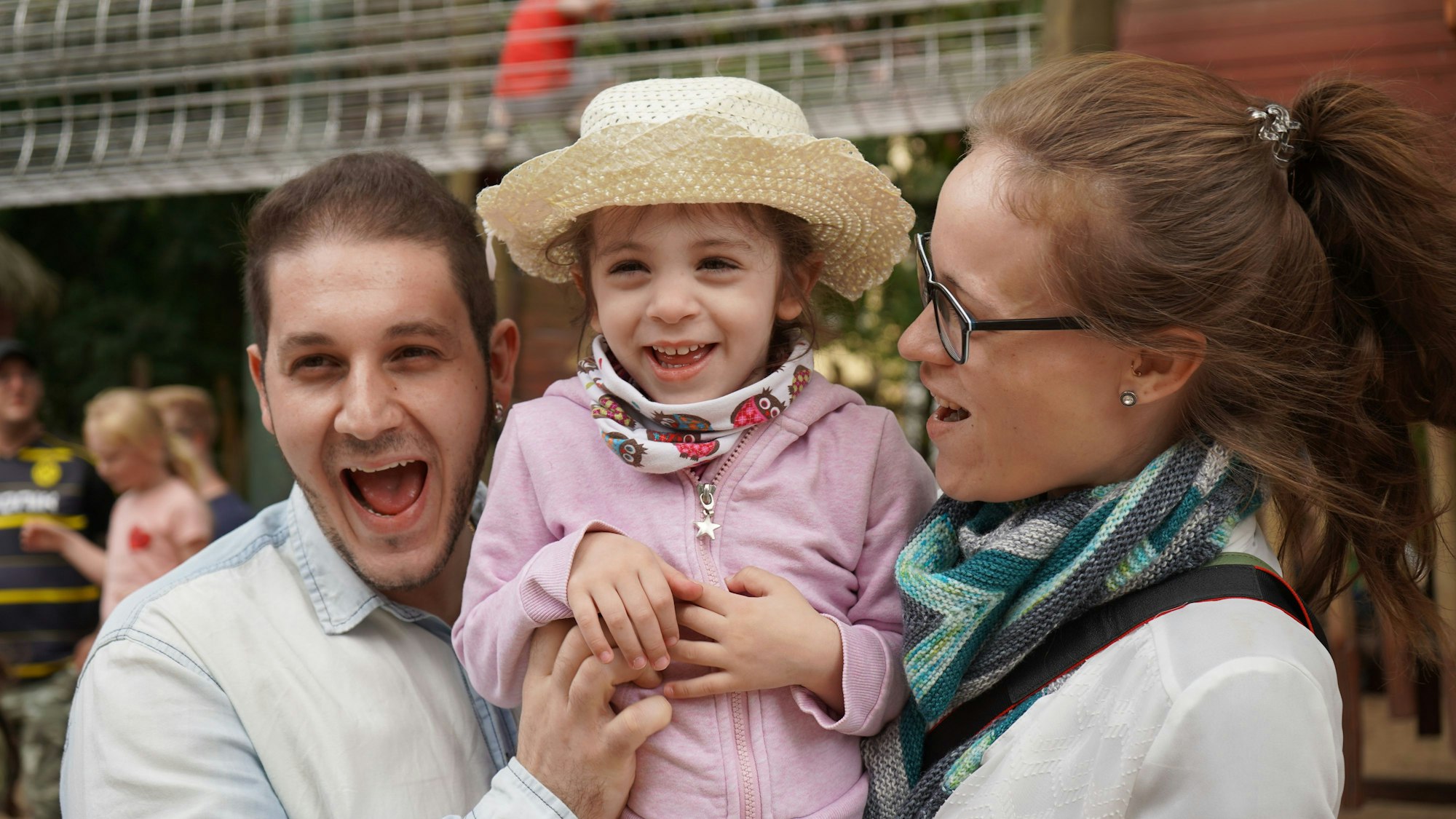 Sofias Eltern Enzo und Jacqueline halten ihre Tochter zwischen sich hoch und lachen. Sofia trägt einen Strohhut.