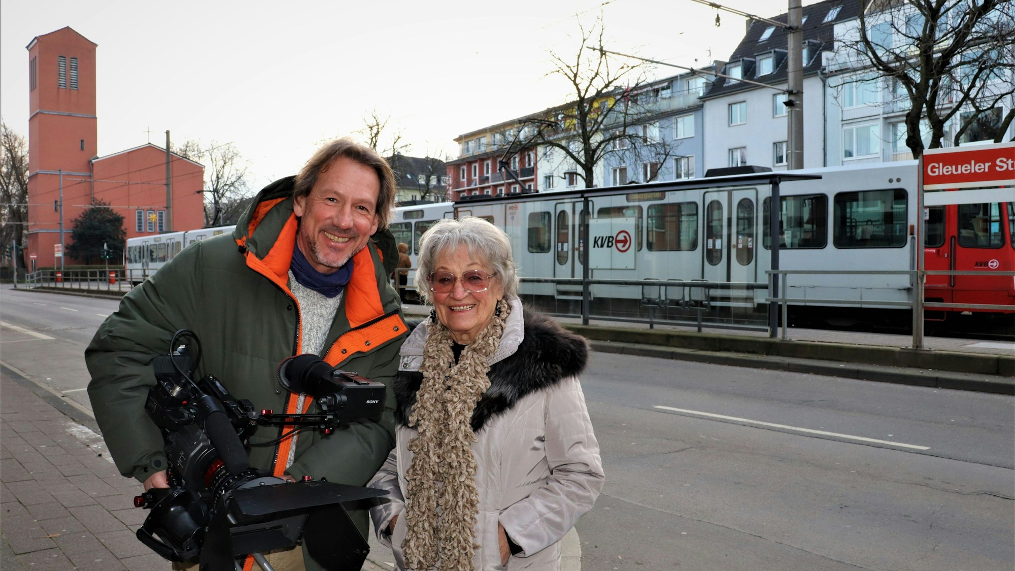 Ein junger Mann mit braunem Haar hält eine Filmkamera, neben ihm steht eine grauhaarige Dame mit Brille und Schal.