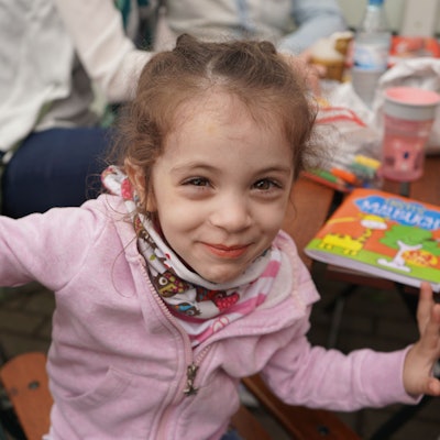 Sofia lächelt und sitzt an auf einem Holzstuhl. Auf dem Tisch neben ihr liegt ein Malbuch.