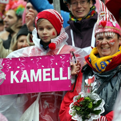 08.02.2016: Rosenmontag in Köln: Jecke stehen verkleidet am Straßenrand und schauen auf den Rosenmontagszug. Ein Kind hält ein Schild, auf dem „Kamelle“ steht.