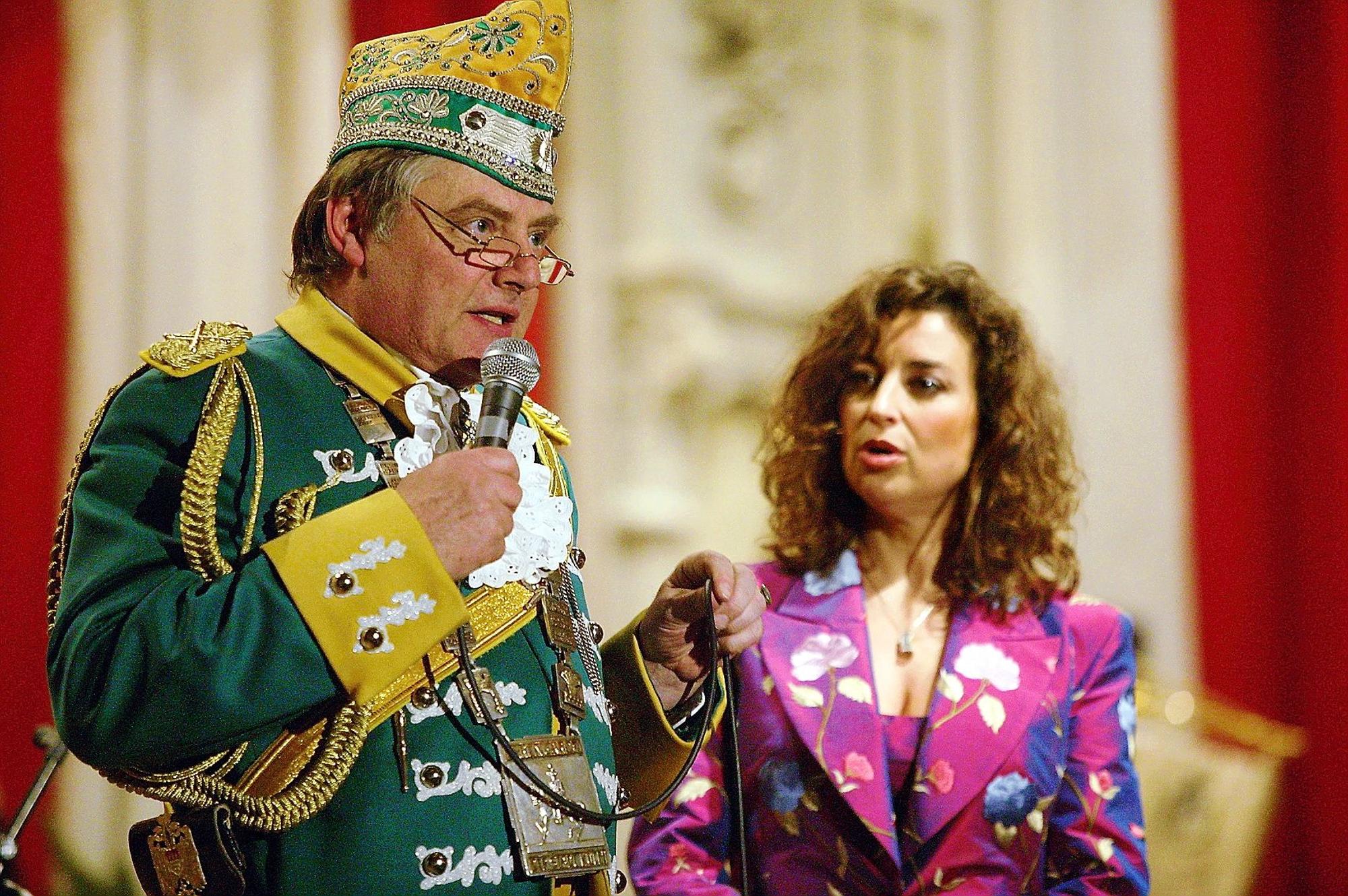 Reiner Benner spricht ins Mikrofon zum Saal, im Hintergrund die konsternierte Isabel Varell.