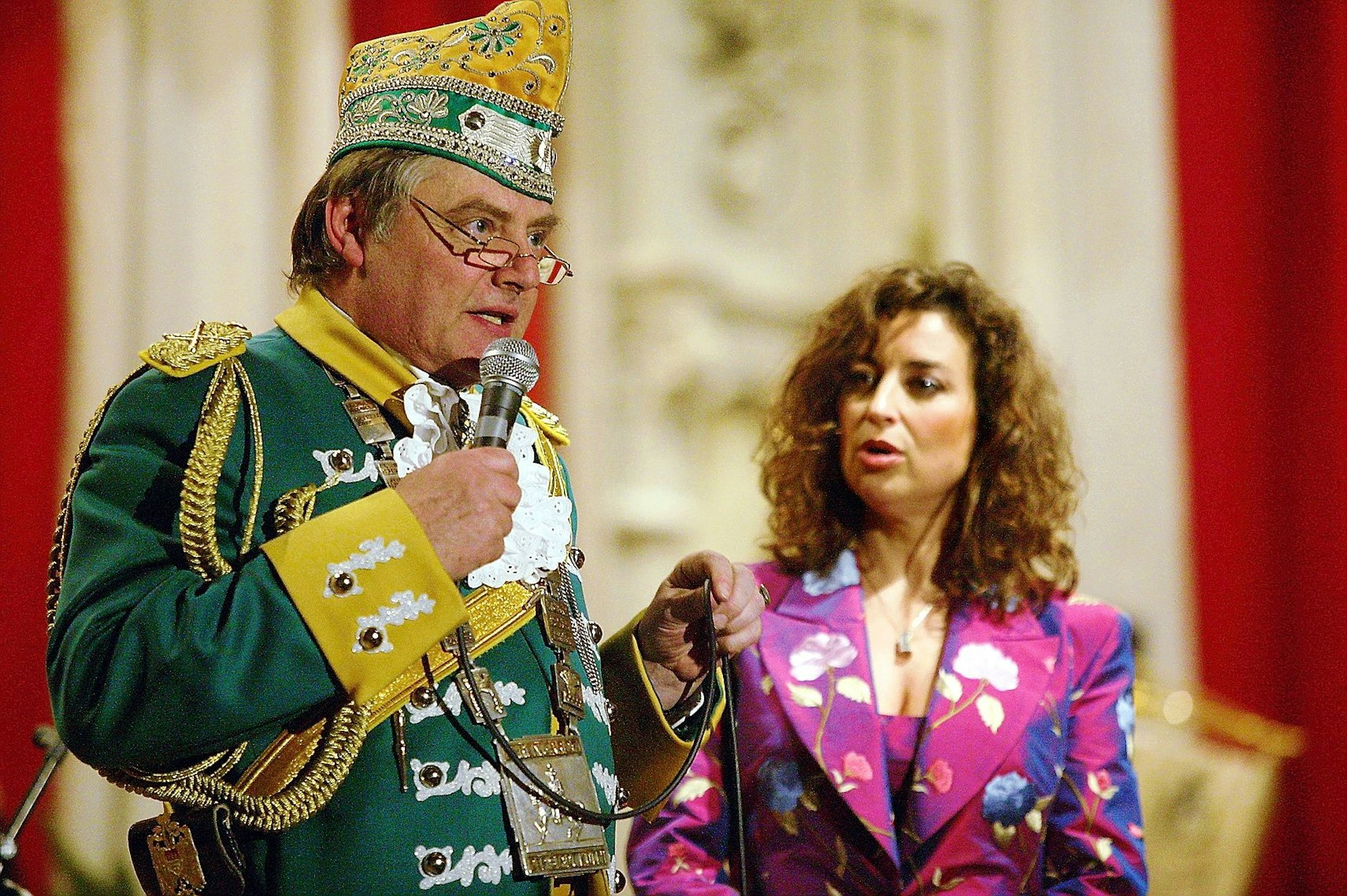 Reiner Benner spricht ins Mikrofon zum Saal, im Hintergrund die konsternierte Isabel Varell.