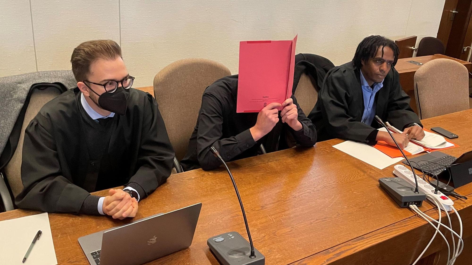 Der Angeklagte sitzt mit einer Mappe vor dem Gesicht zwischen seinen Verteidigern Maximilian Klefenz (l.) und Dawit Stefanos beim Prozessauftakt im Landgericht.