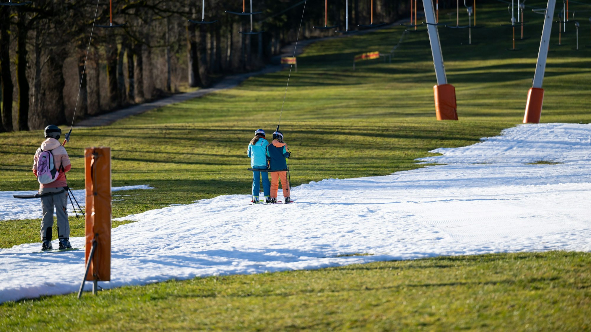 Wintersportler fahren in einem bayrischen Skigebiet zwischen grünen Wiesen mit einem Schlepplift die beschneite Piste hinauf. Auch die Natur leidet unter der ungewöhnlichen Wärme.