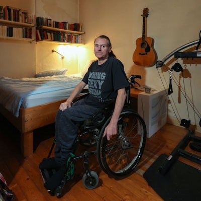 Ralf Degen sitzt im Rollstuhl. Hinter ihm ist sein Bett zu sehen, rechts von ihm ein Fitnessgerät. Er trägt ein T-Shirt mit der Aufschrift Young, Sexy, Muscican.