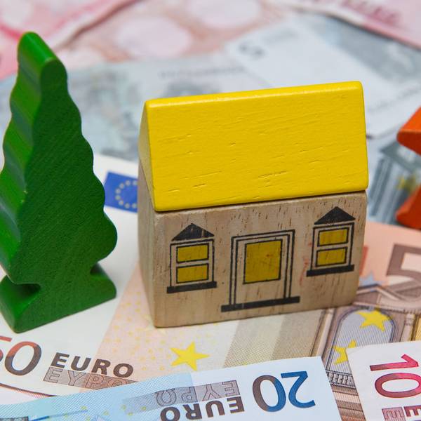 Man sieht ein Bauklotz-Haus, das auf Geldscheinen steht. Rechts neben dem Haus steht eine Holzfigur, links ein Holzbaum.