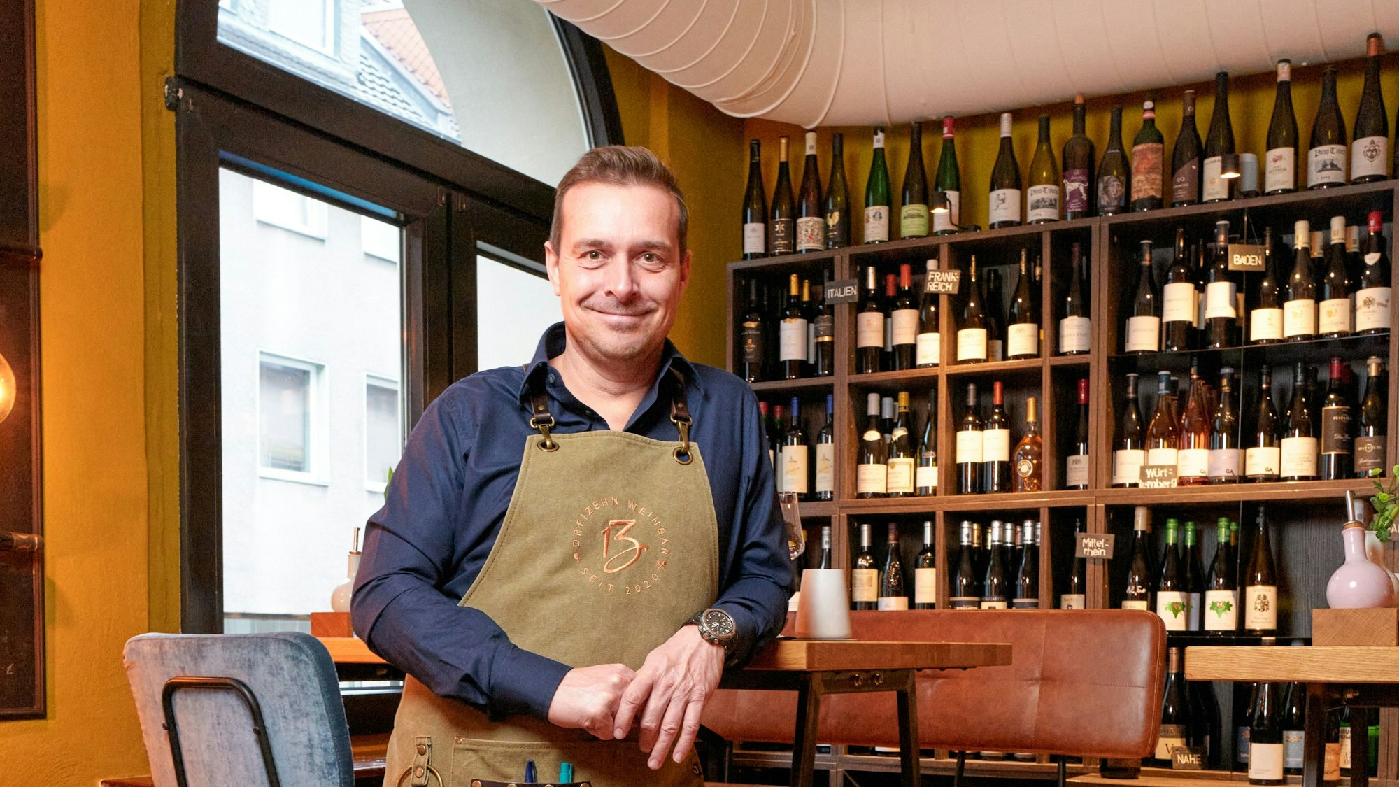 Mann mit Schürze steht vor einem Regal mit Weinflaschen