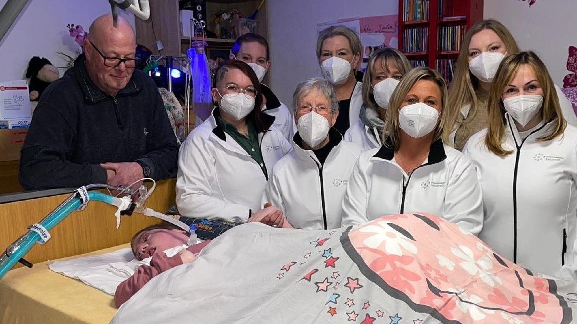 Man sieht Julia in einem Pflegebett liegen, am Kopfende ihr Vater, daneben acht Personen des Pflegeteams.