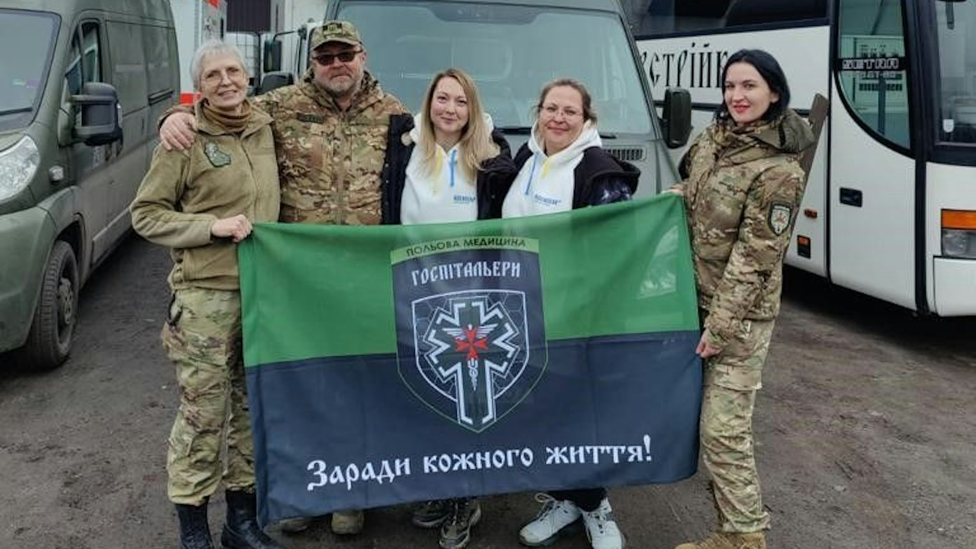 Vier Frauen und ein Mann, teilweise in Militärkleidung, stehen mit einer ukrainischen Fahne vor einem olivgrün gefärbten Transporter.