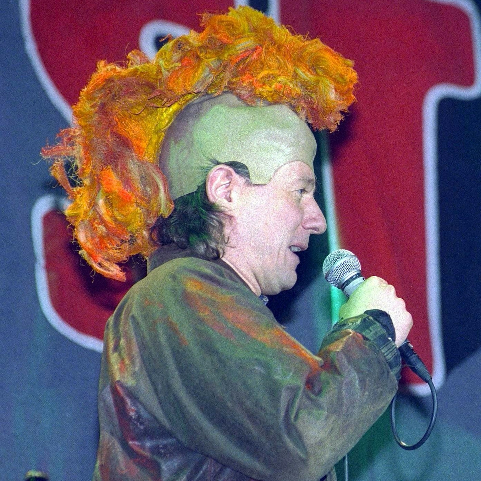 Jürgen Becker hat eine Irokesen-Perücke auf und spricht ins Mikrofon.