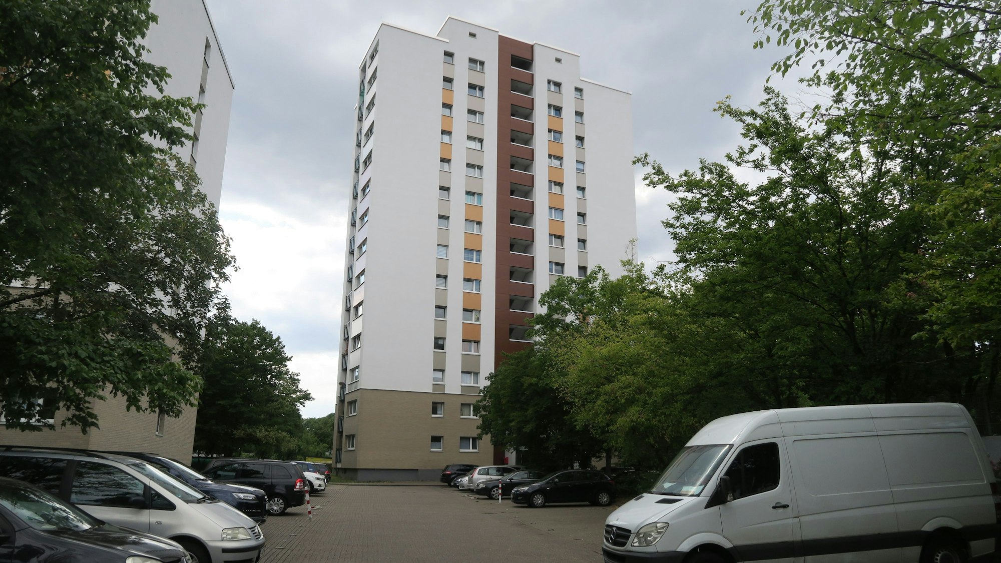 Ein Hochhaus ist zu sehen, davor ein Parkplatz mit einigen Autos vor sommerlich grünen Bäumen.