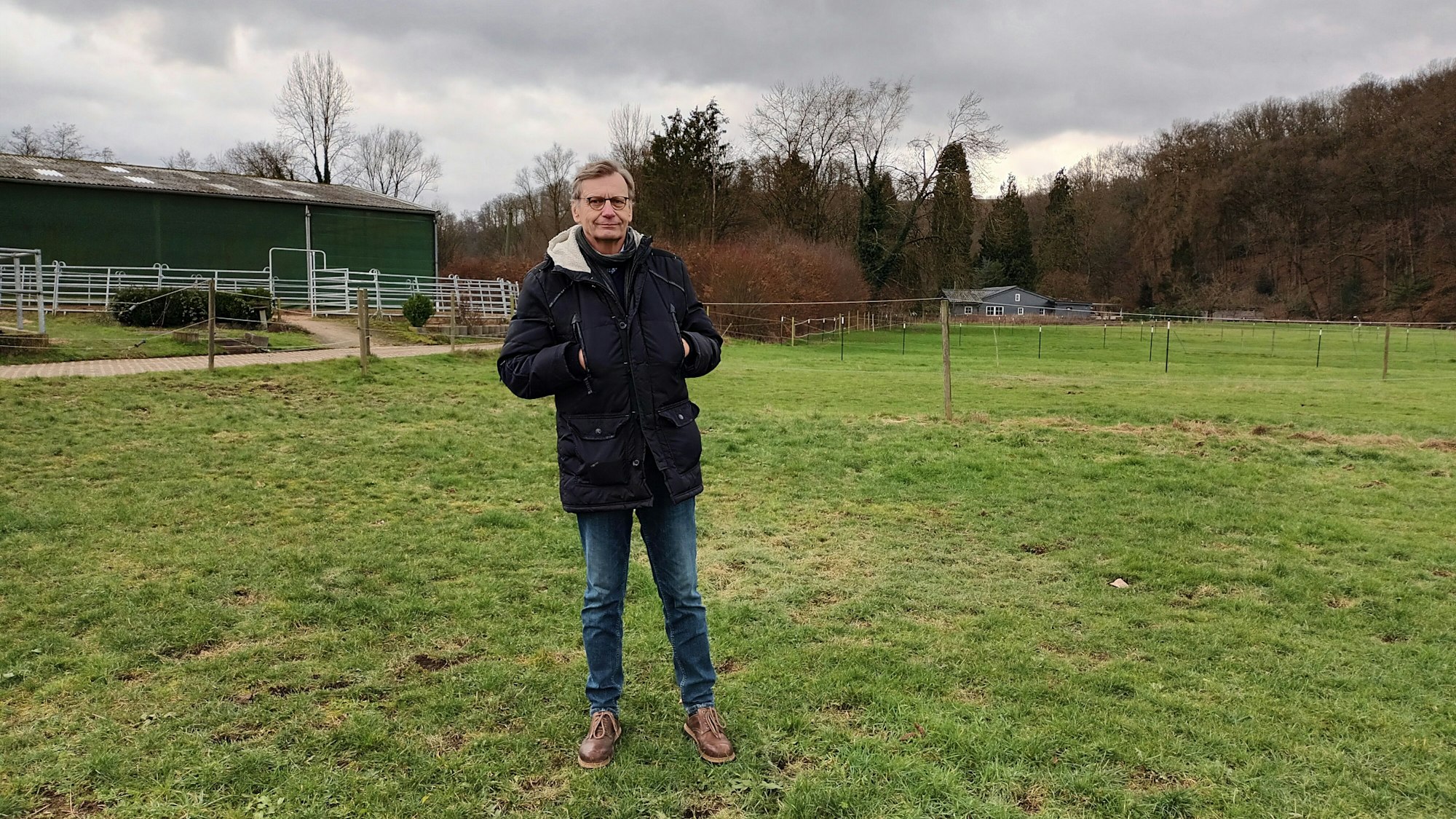 Ein Mann im Anorak steht auf einer grünen Wiese. Im Hintergrund steht ein Pferdestall.