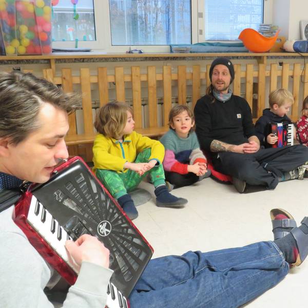 Kinder und zwei Erzieher sitzen auf dem Boden und singen gemeinsam. Ein Erzieher spielt auf einem Akkordeon.