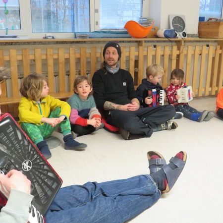 Kinder und zwei Erzieher sitzen auf dem Boden und singen gemeinsam. Ein Erzieher spielt auf einem Akkordeon.