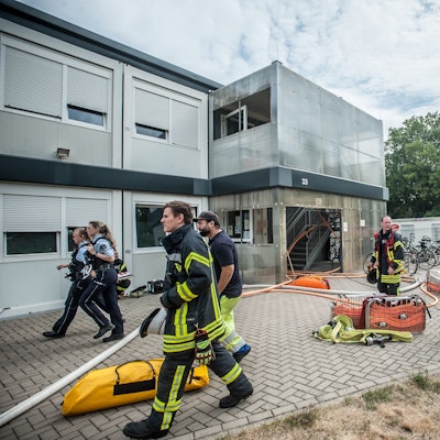 Feuerwehrleute stehen vor der Containersiedlung für Flüchtlinge in der Heinrich-Claes-Straße 33