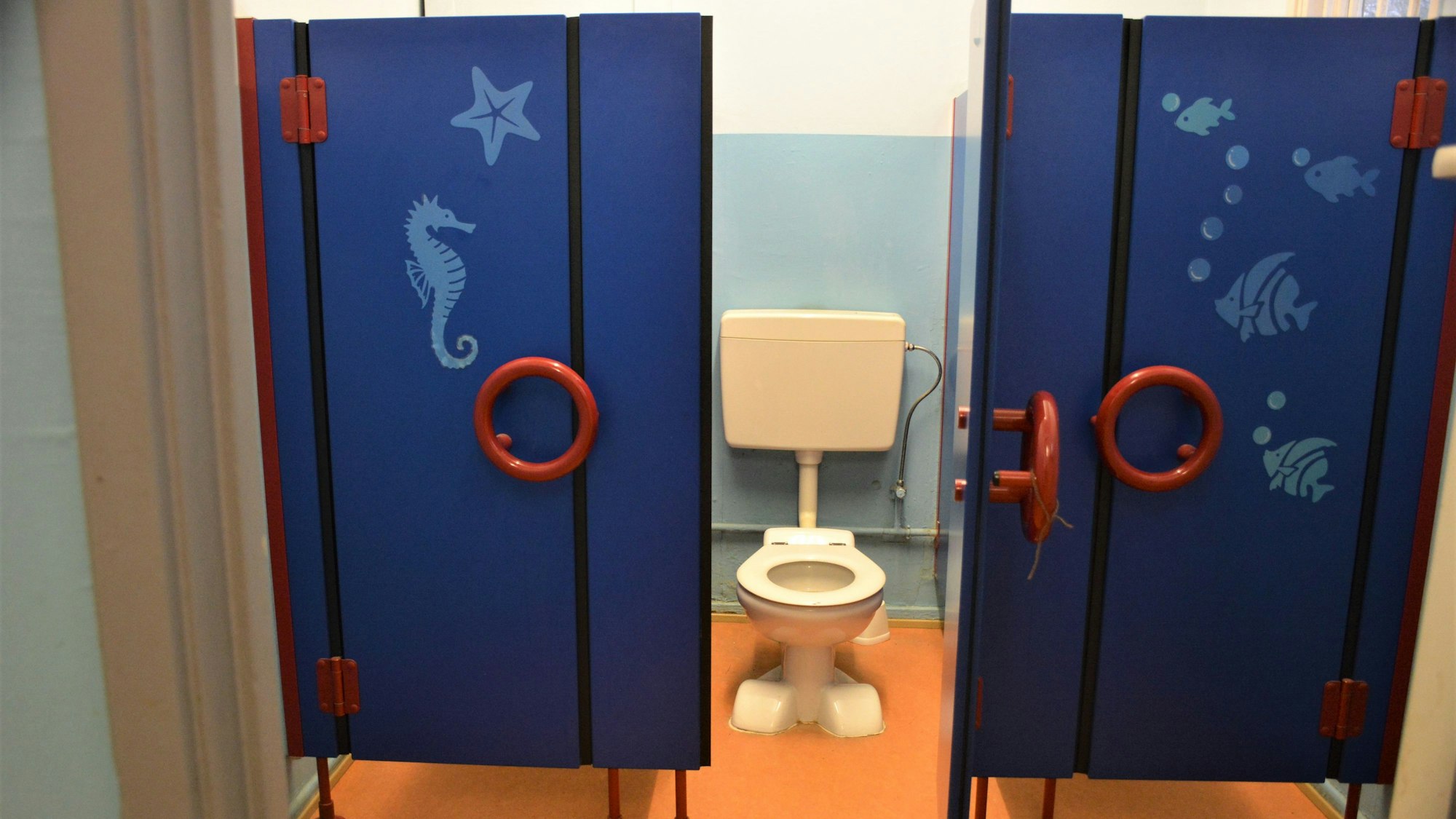 Zu sehen sind zwei blaue Toilettentüren mit Seepferdchen-Motiven und eine sehr kleine Kloschüssel.