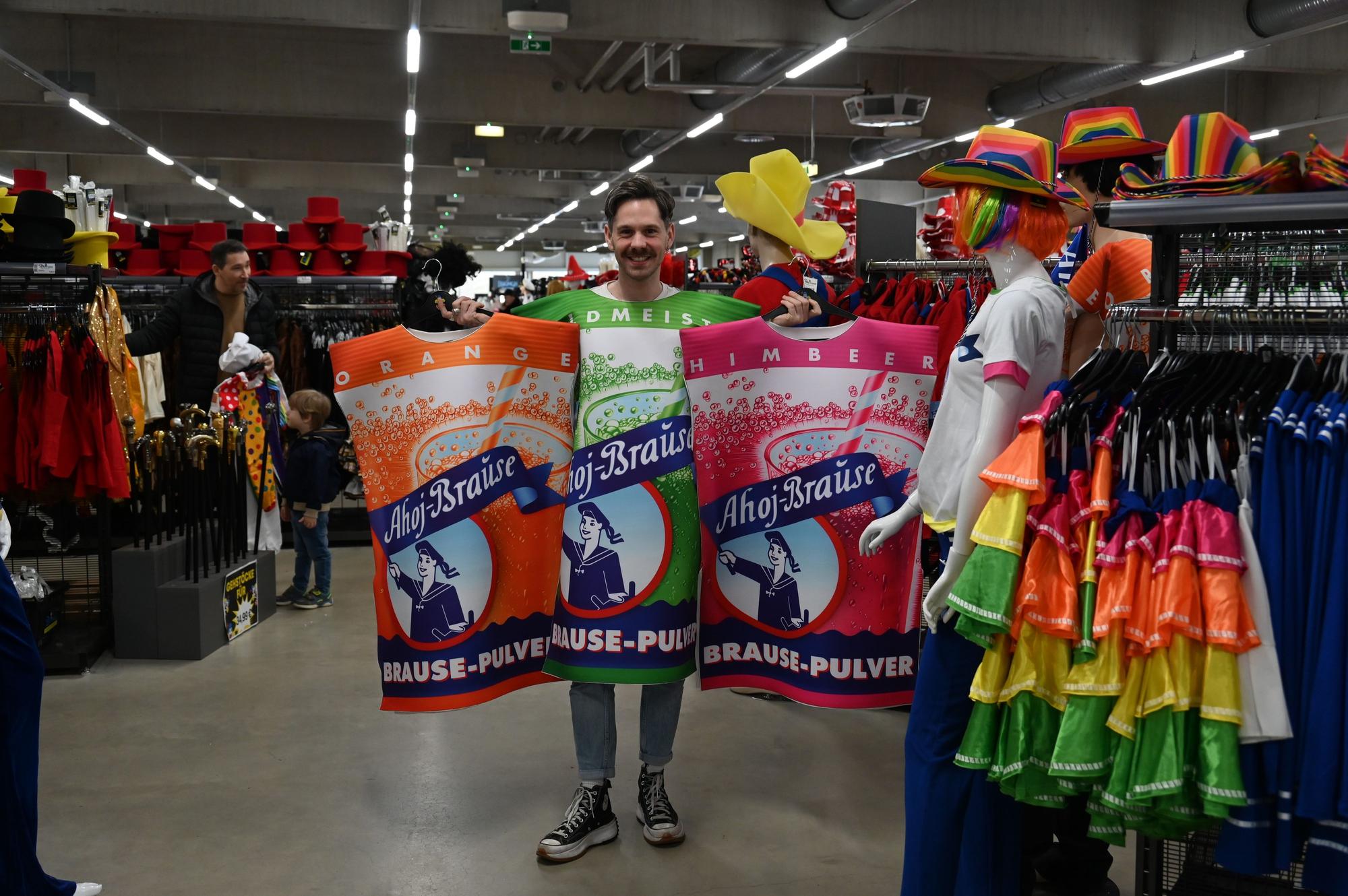 Ein Mann ist als grüne Ahoj-Brause verkleidet und hält zwei weitere Ahoj-Brause-Kostüme in einem Karnevalsladen hoch.