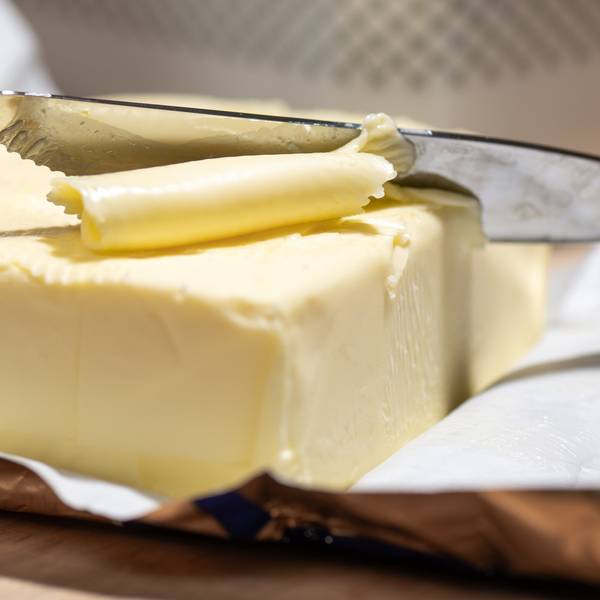 Ein Stück Butter liegt in der Verpackung auf dem Tisch, jemand nimmt mit dem Messer davon.