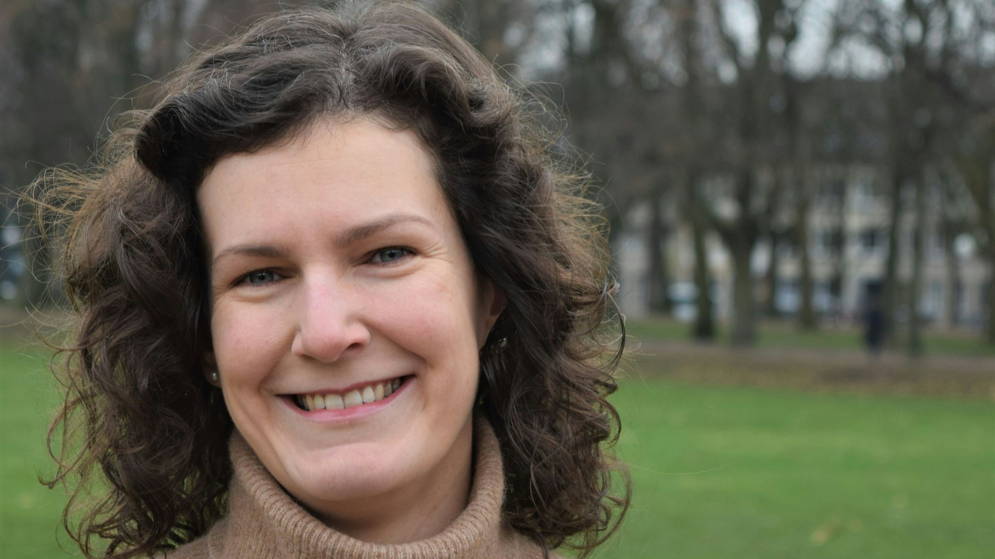 Dagny Schwarz schaut für ein Porträt in die Kamera. Sie trägt ein braunes Winterkleid mit Rollkragen und hat schulterlange, lockige braune Haare.