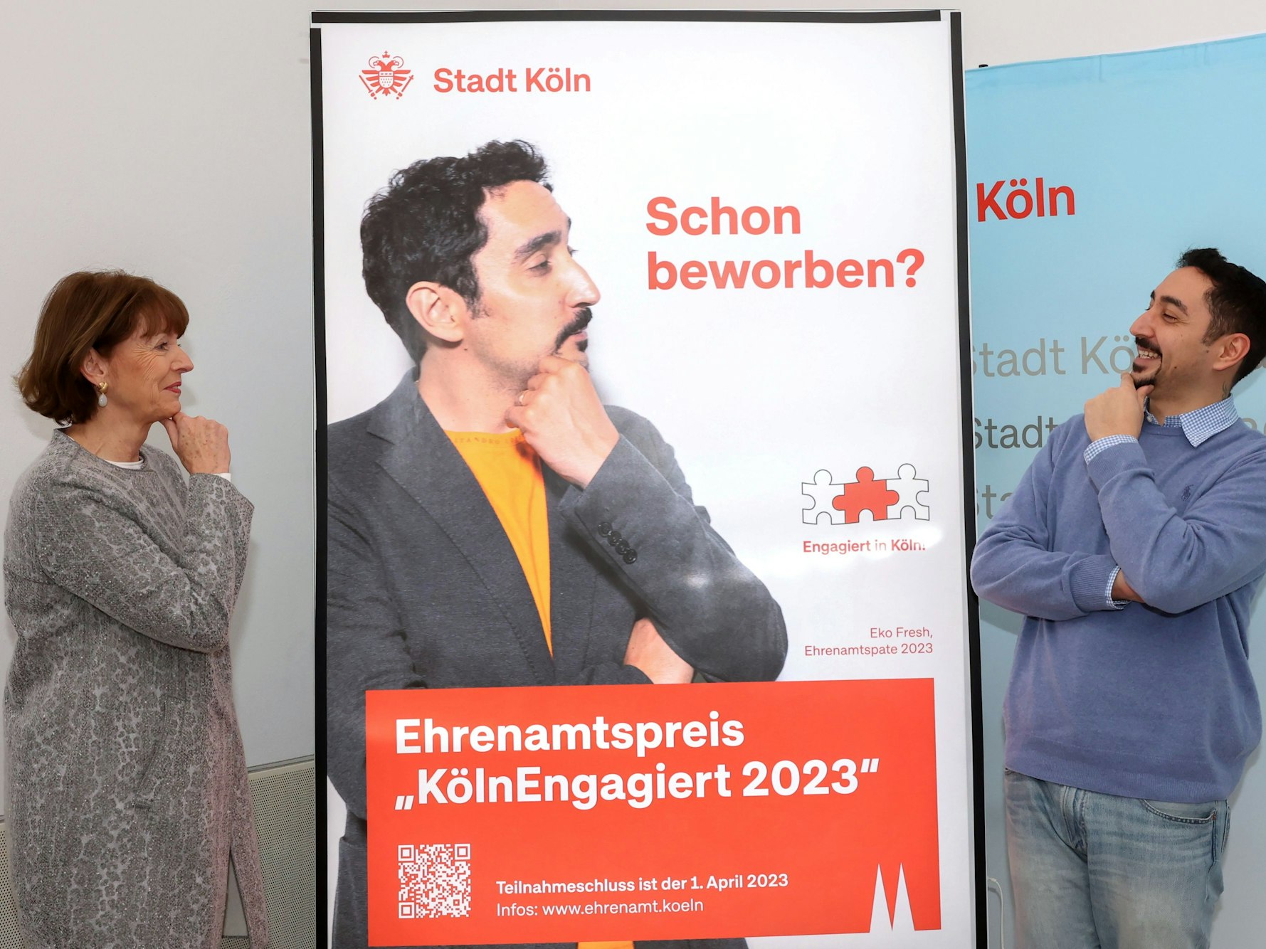 OB Reker und Rapper Eko Fresh posieren vor einem Werbeplakat für den Kölner Ehrenamtspreis.