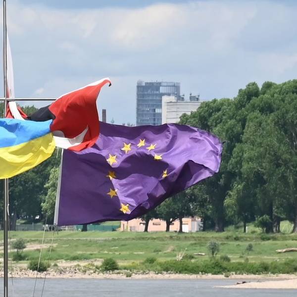 Eine ukrainische Fahne weht neben einer EU-Fahne im Wind, im Hintergrund der Rhein.
