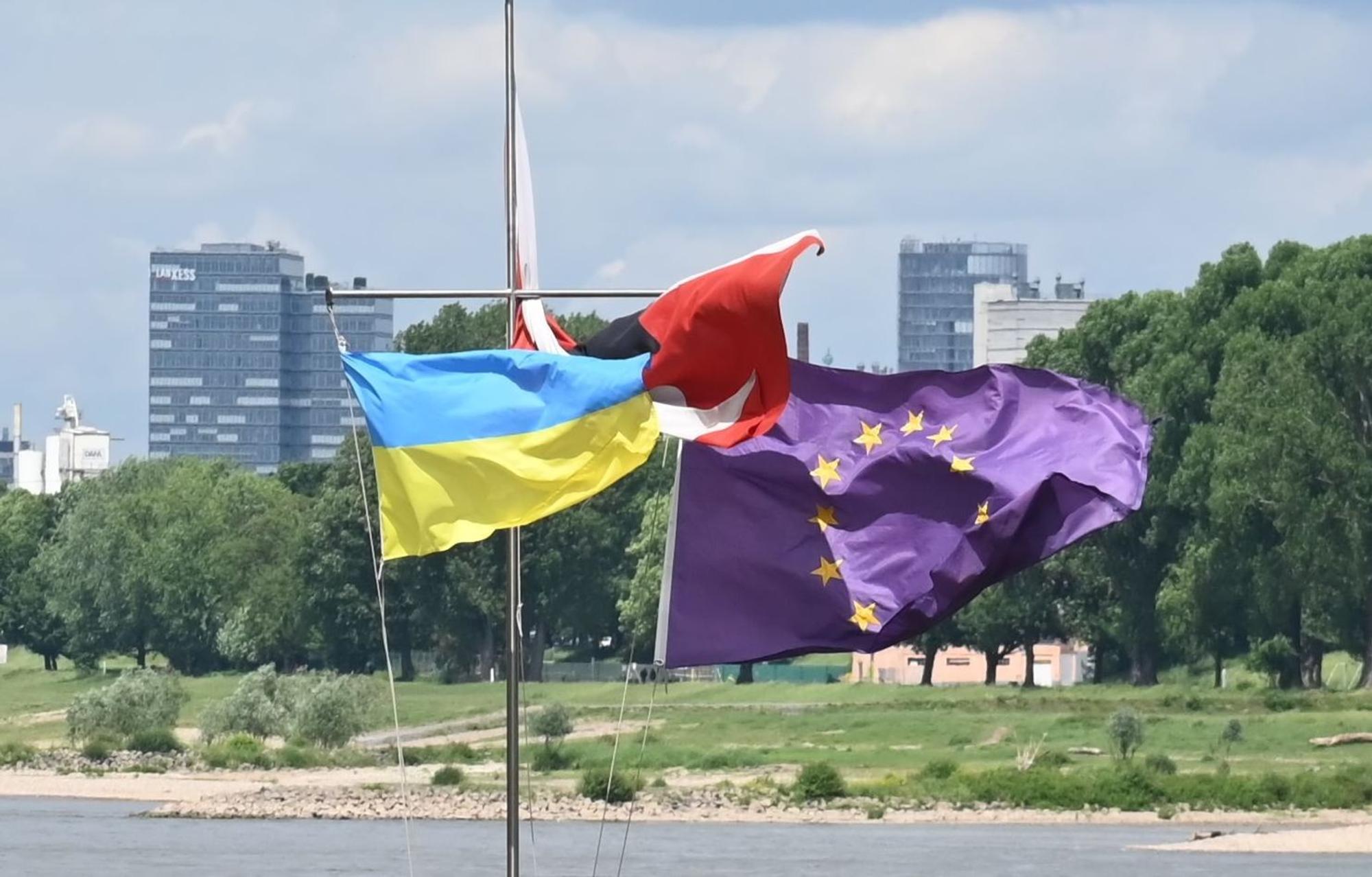 Eine ukrainische Fahne weht neben einer EU-Fahne im Wind, im Hintergrund der Rhein.